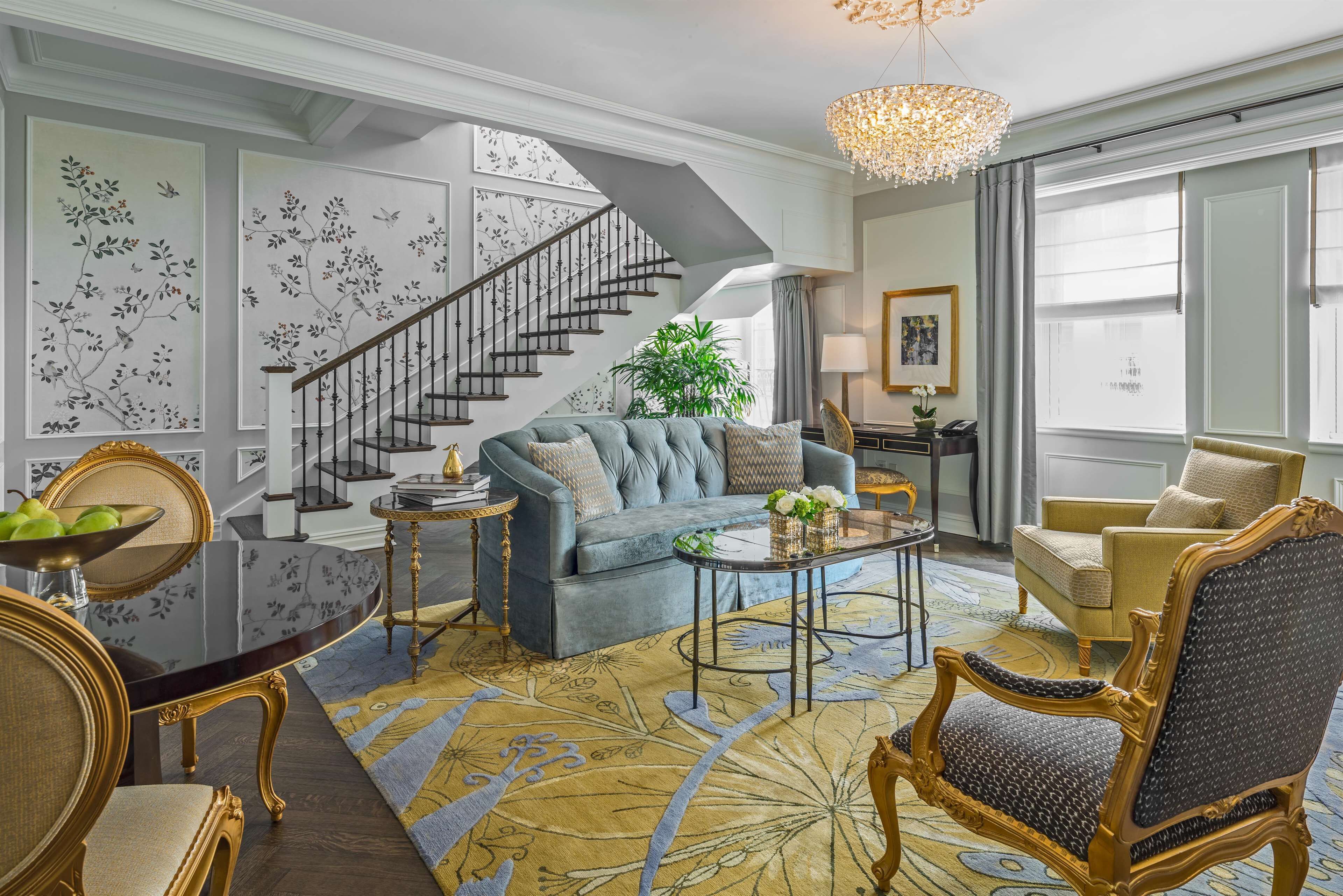 A spacious living room with a blue sofa, a dining table with chairs, a staircase, and a large window with curtains.