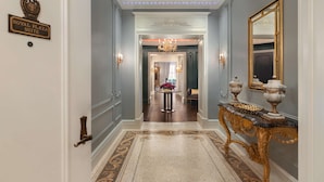 A hallway with a sign reading "ROYAL PLAZA SUITE," a chandelier, a gold console table with vases, and a patterned carpet.