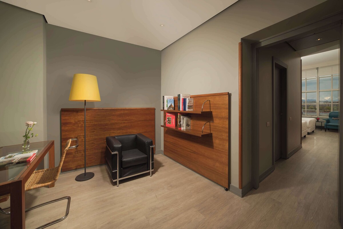 Un moderno vestíbulo de hotel con un mostrador de recepción de madera, una butaca de cuero negro, una mesa de vidrio con un jarrón de flores y una lámpara de pie.