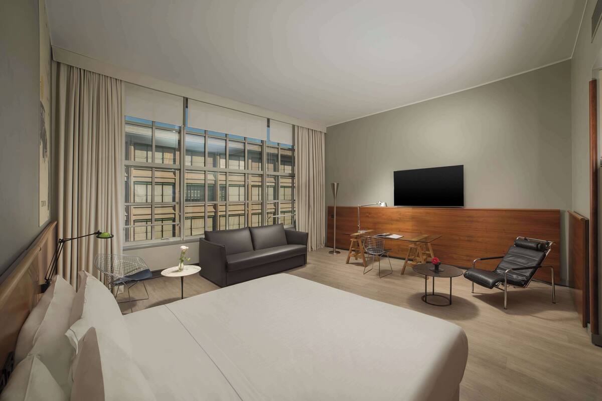 Una habitació d'hotel amb un llit gran, un sofà, una butaca, una taula petita, un televisor fixat a la paret i una finestra amb cortines.