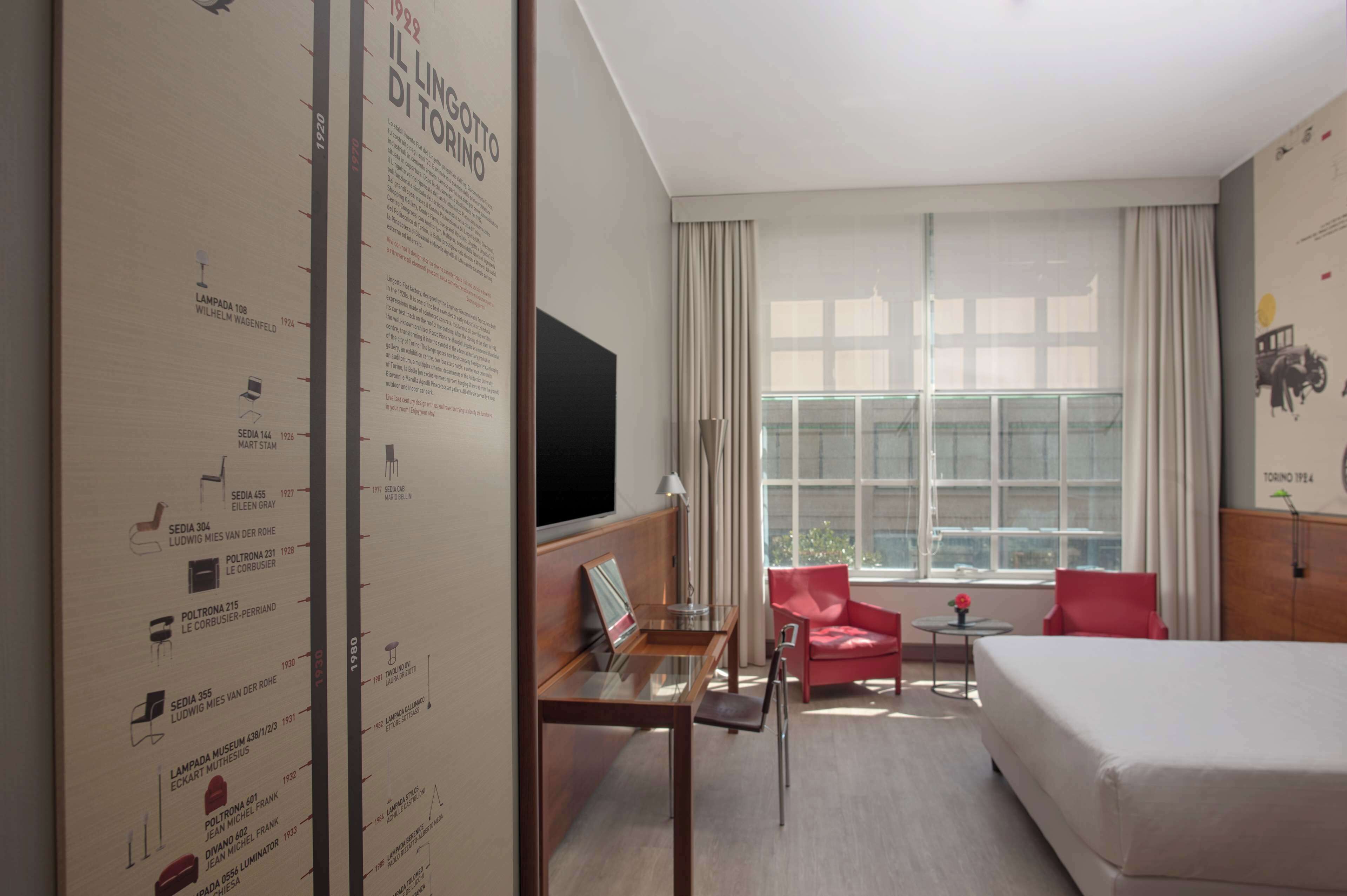 Quarto de hotel com cama, escrivaninha com cadeira, televisão e uma grande janela com cortinas.
