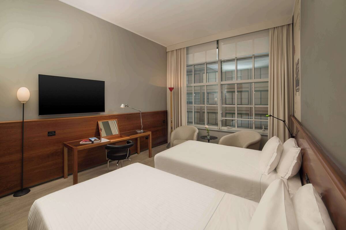 A hotel room with two beds, a desk with a chair, a TV, and a large window with curtains.