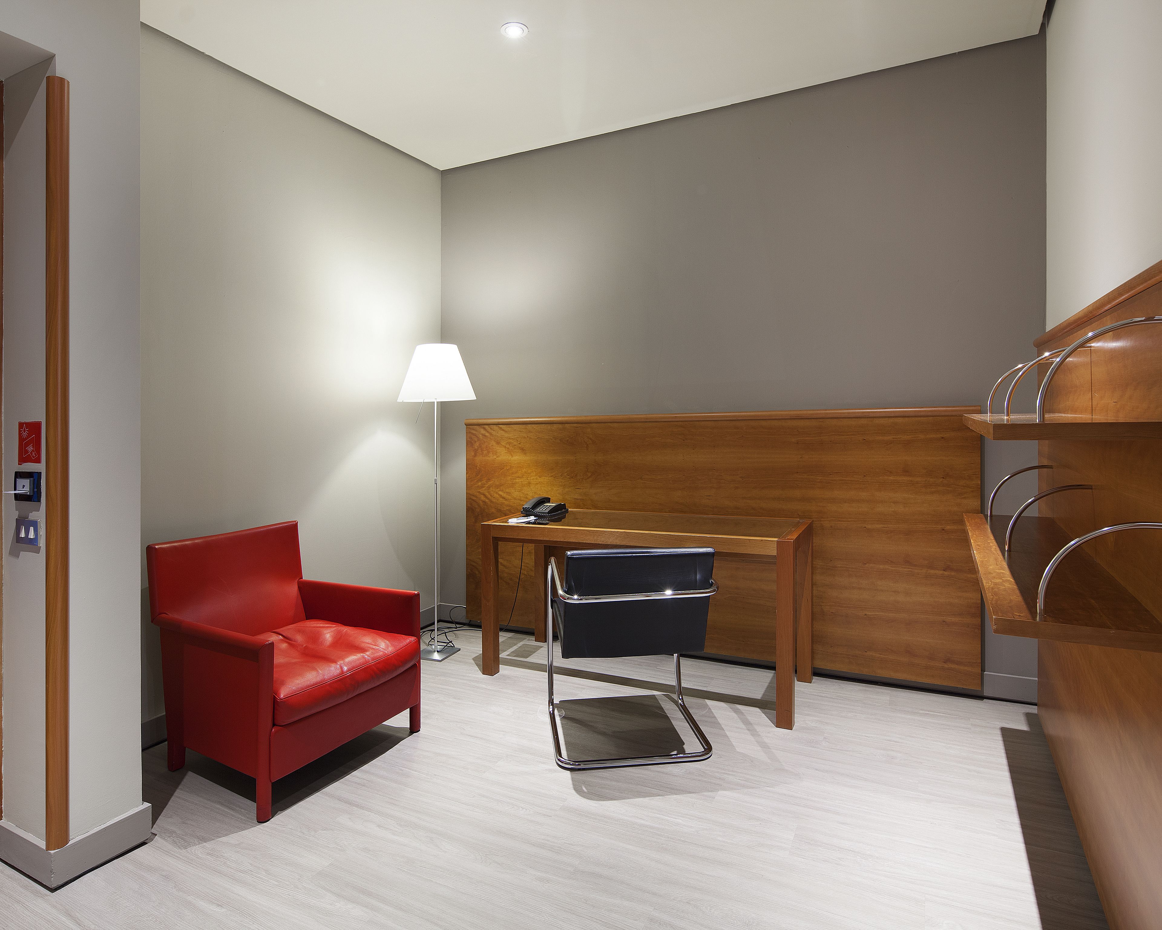 A modern hotel room with a red chair, a desk with a lamp, and a wooden headboard.