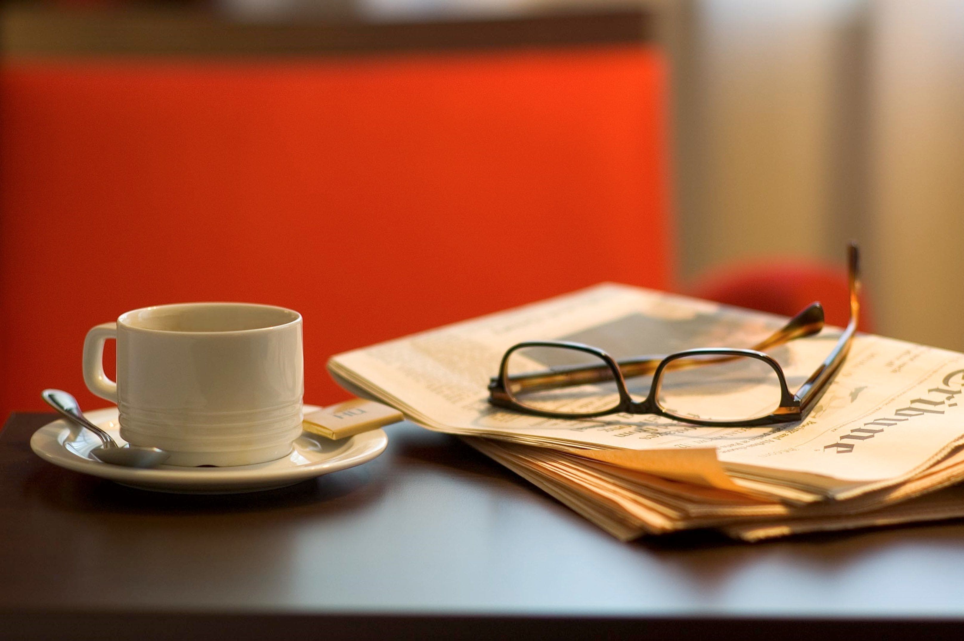 A white cup on a saucer with a spoon, glasses, and newspapers on a table.