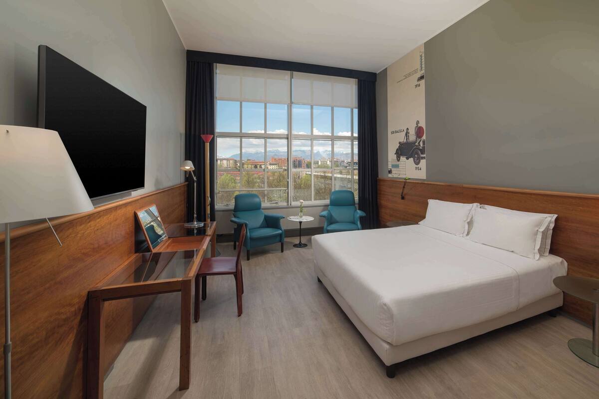 A hotel room with a large bed, a desk with a chair, a small table with two chairs, and a large window offering a city view.