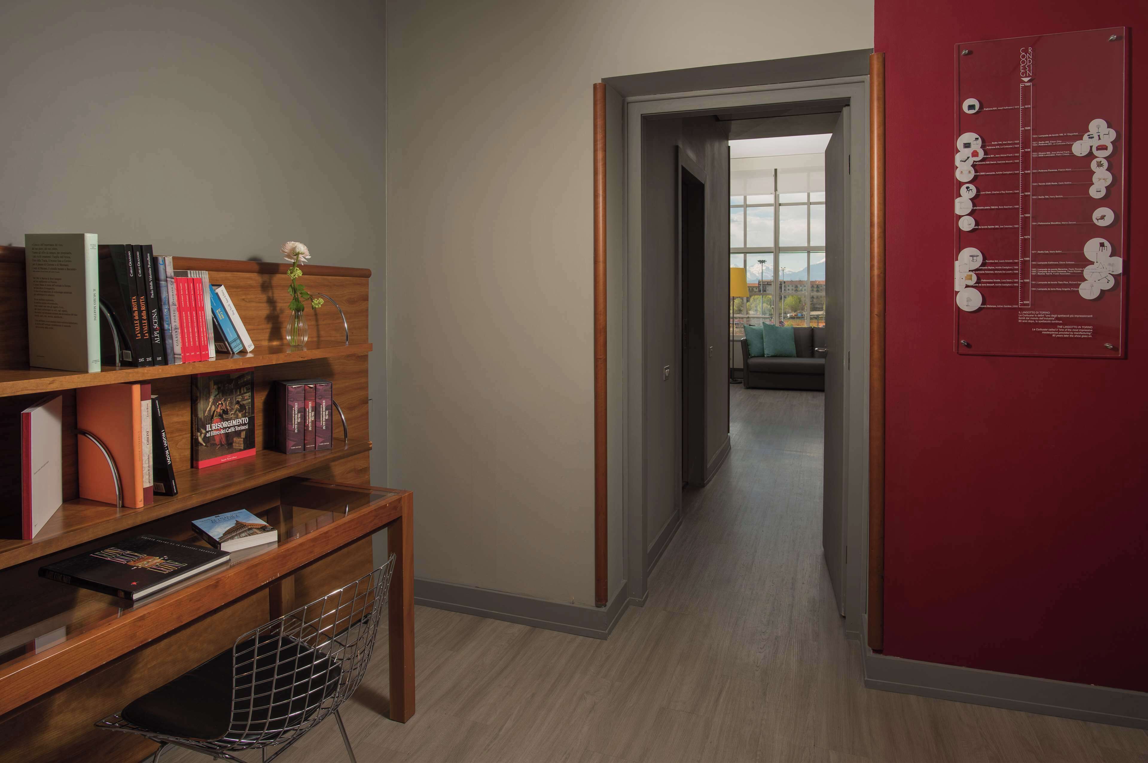 A hallway with a wooden bookshelf, a red bulletin board, and a window leading to another room.