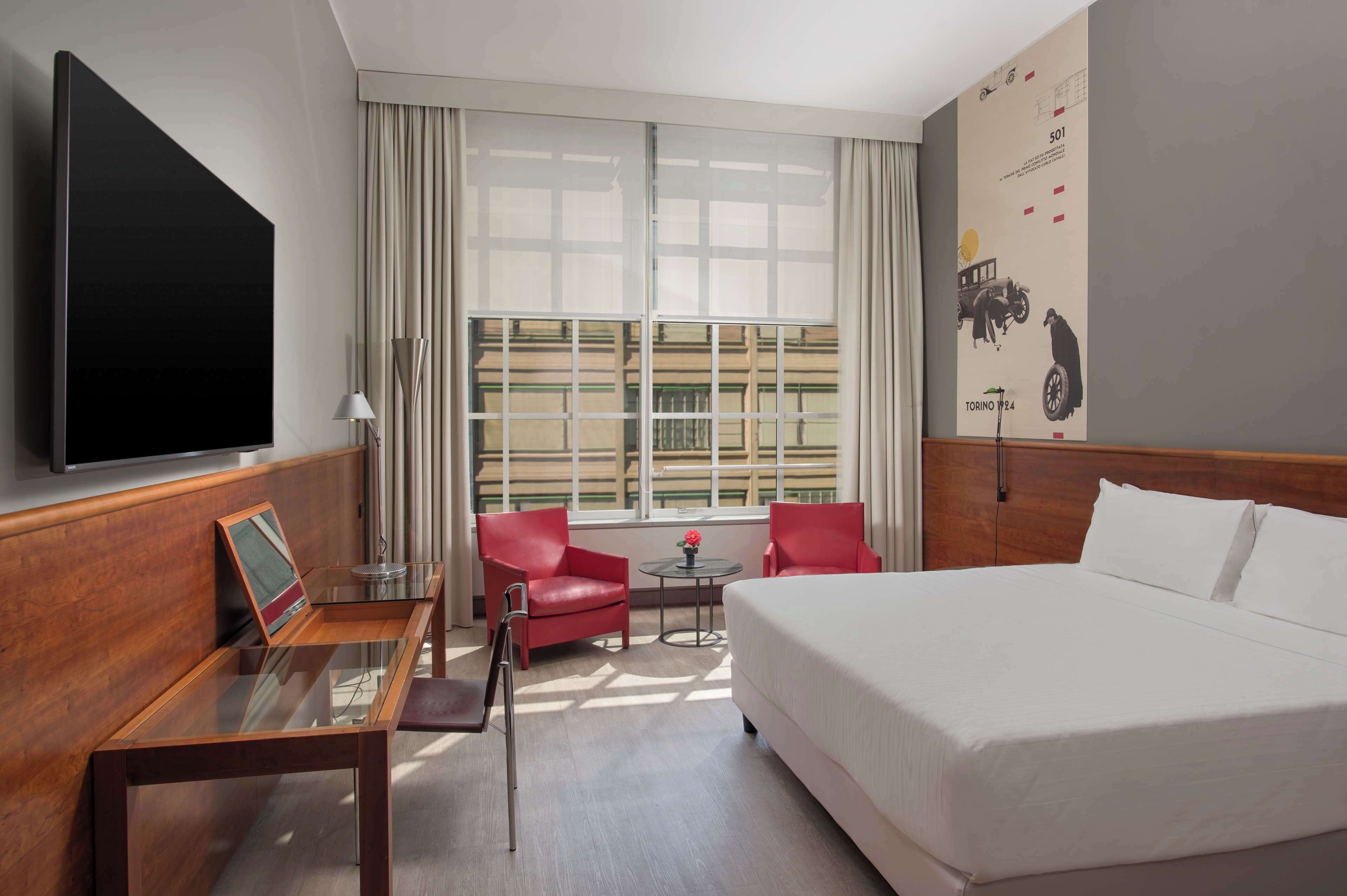 A hotel room with a large bed, a TV, a desk with a chair, a small table, and two red chairs.
