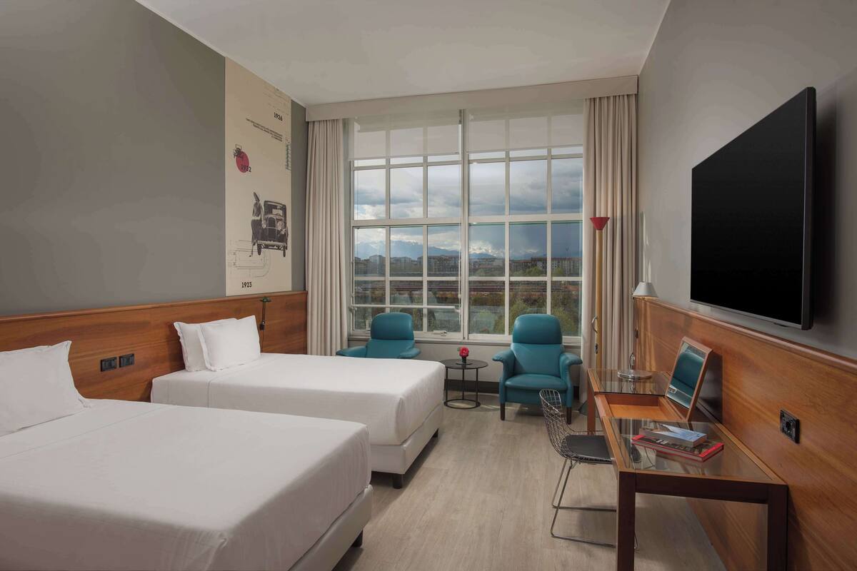 Habitació d'hotel amb dos llits, televisió, taula petita amb una cadira i finestra amb vista a edificis i muntanyes.