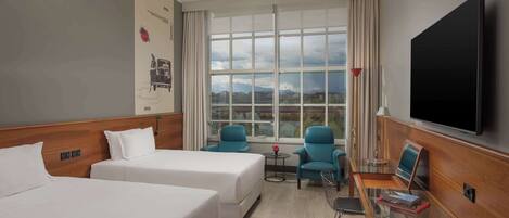 Habitación de hotel con dos camas, televisión, una mesita con una silla y ventana con vistas a edificios y montañas.