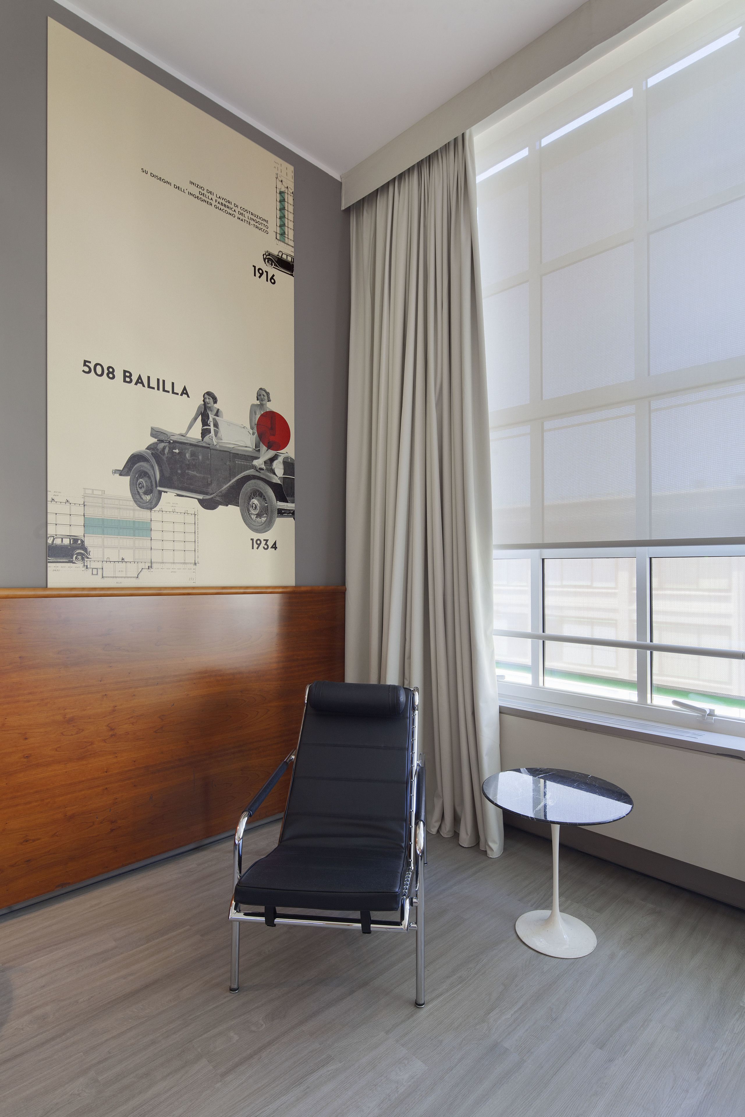 Una camera d'albergo moderna con parete rivestita in legno, una poltrona lounge nera, un tavolino rotondo e una grande finestra con tende.