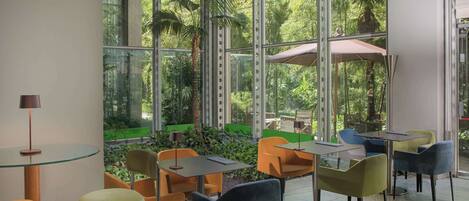 A modern indoor dining area with glass walls, multiple tables and chairs, and a view of a lush garden.