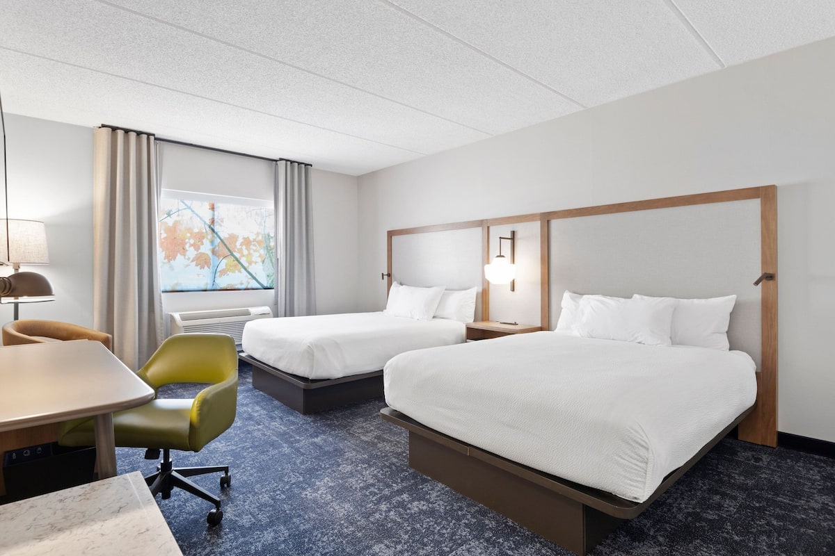 A hotel room with two beds, a desk, a chair, a lamp, and a window with a view of trees.