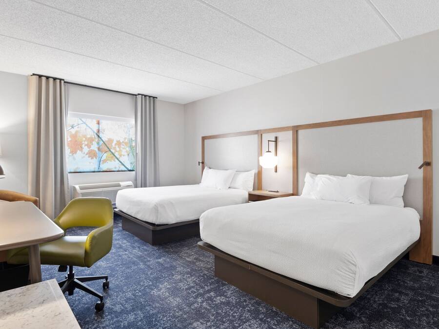A hotel room with two beds, a desk, a chair, a lamp, and a window with a view of trees.