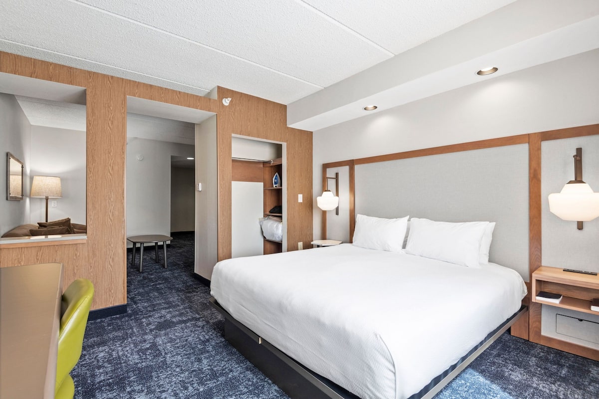 A hotel room with a large bed, a desk with a lamp, a chair, and a wardrobe.