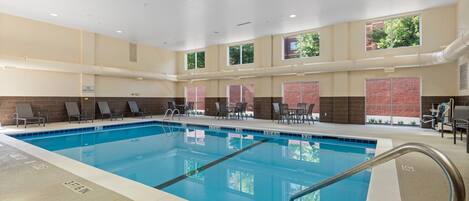 A large indoor swimming pool with clear lane markings, lounge chairs, and a view of a brick building through the windows.
