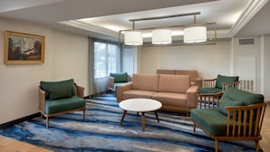 Une chambre d’hôtel moderne avec un canapé beige, deux fauteuils verts, une table basse ronde et une grande fenêtre avec des rideaux.