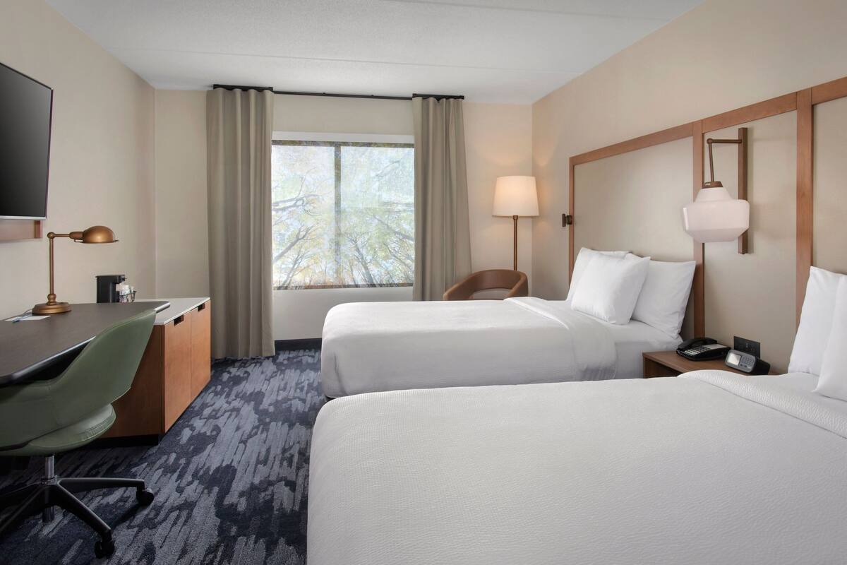 A hotel room with two beds, a desk, a chair, a TV, and a window with curtains.
