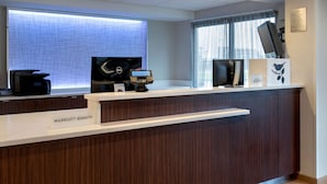 Un comptoir d’accueil avec une plaque nominative indiquant « MARRIOTT BONVOY », un écran numérique, une machine à café et une fenêtre avec des rideaux.