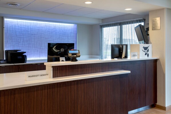 A reception desk with a nameplate reading "MARRIOTT BONVOY," a digital screen, a coffee machine, and a window with curtains. - Fairfield Inn Marriott Rochester Airport (Rochester)