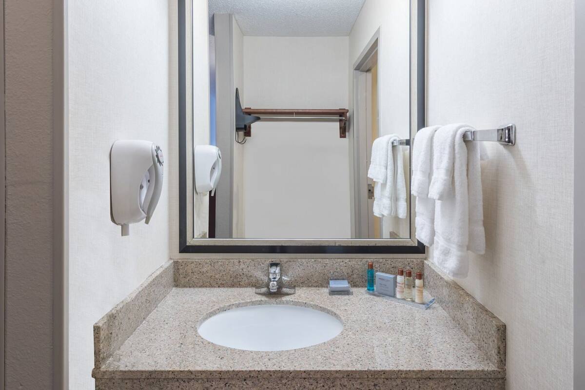 A bathroom with a granite countertop sink, a mirror, a towel rack with white towels, and a hand dryer.