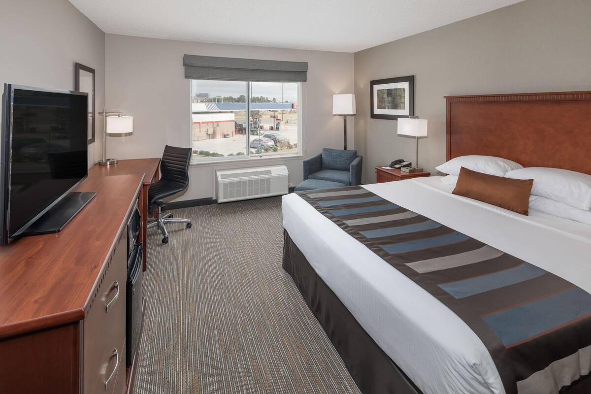 A hotel room with a bed, a desk with a TV, a chair, a nightstand, a lamp, and a window with a view of a building.