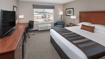 A hotel room with a bed, a desk with a TV, a chair, a nightstand, a lamp, and a window with a view of a building.