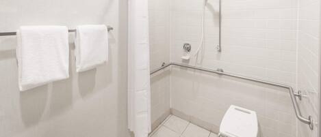 Baño con azulejos blancos, ducha con cabezal de ducha de mano, barra de apoyo, inodoro blanco y dos toallas en un perchero.