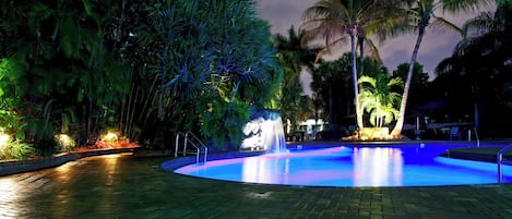 Une piscine nocturne avec une fontaine, entourée de palmiers et illuminée par un éclairage artificiel.