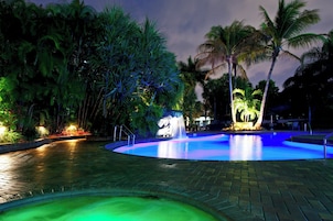 Una piscina iluminada con luces artificiales, con una fuente y rodeada de palmeras, durante la noche.