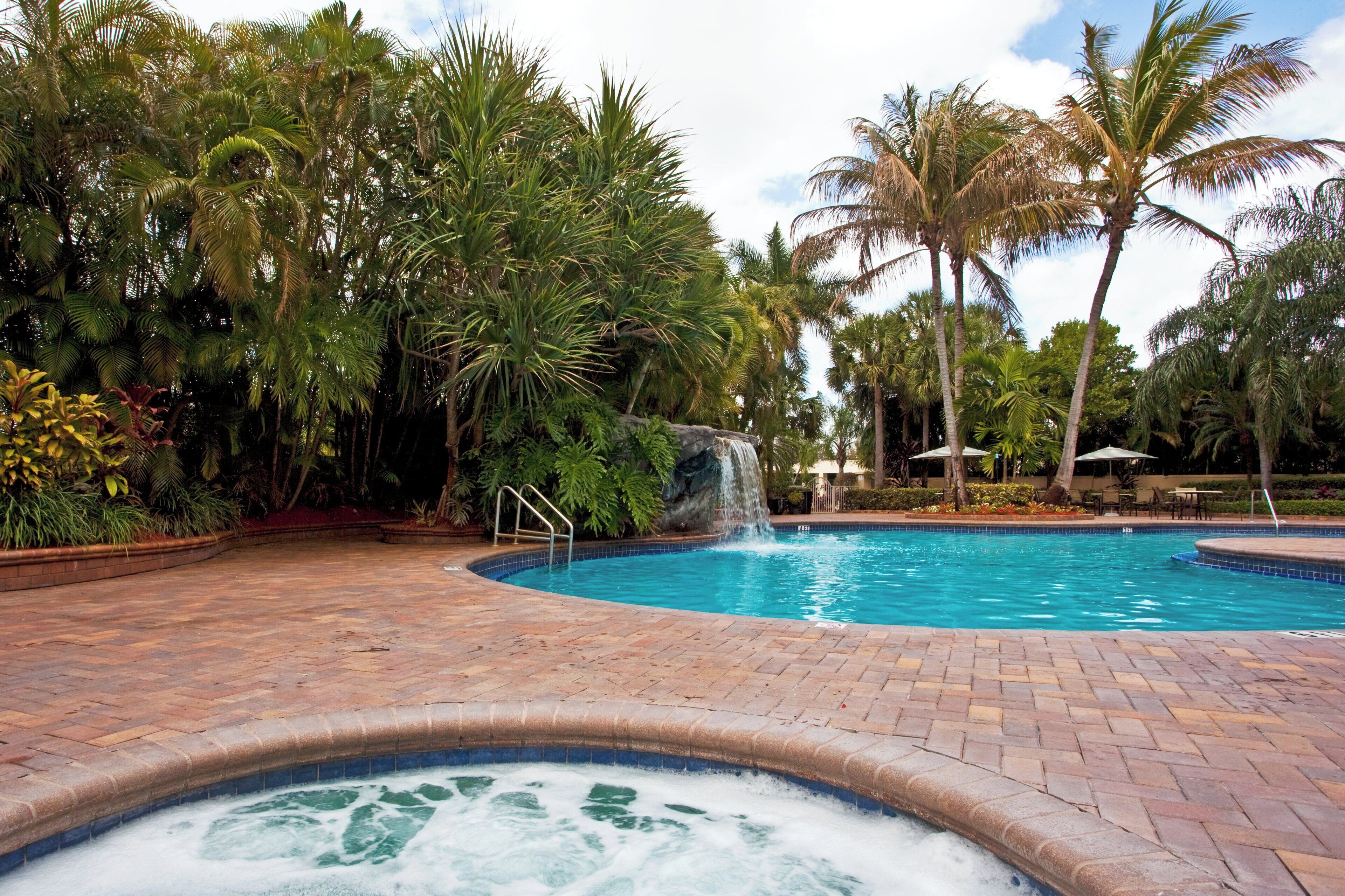 A swimming pool with a hot tub, surrounded by palm trees and a waterfall feature.