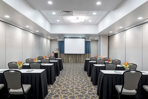 Un salón de conferencias con una pantalla de proyector, mesas con sillas y una alfombra estampada.