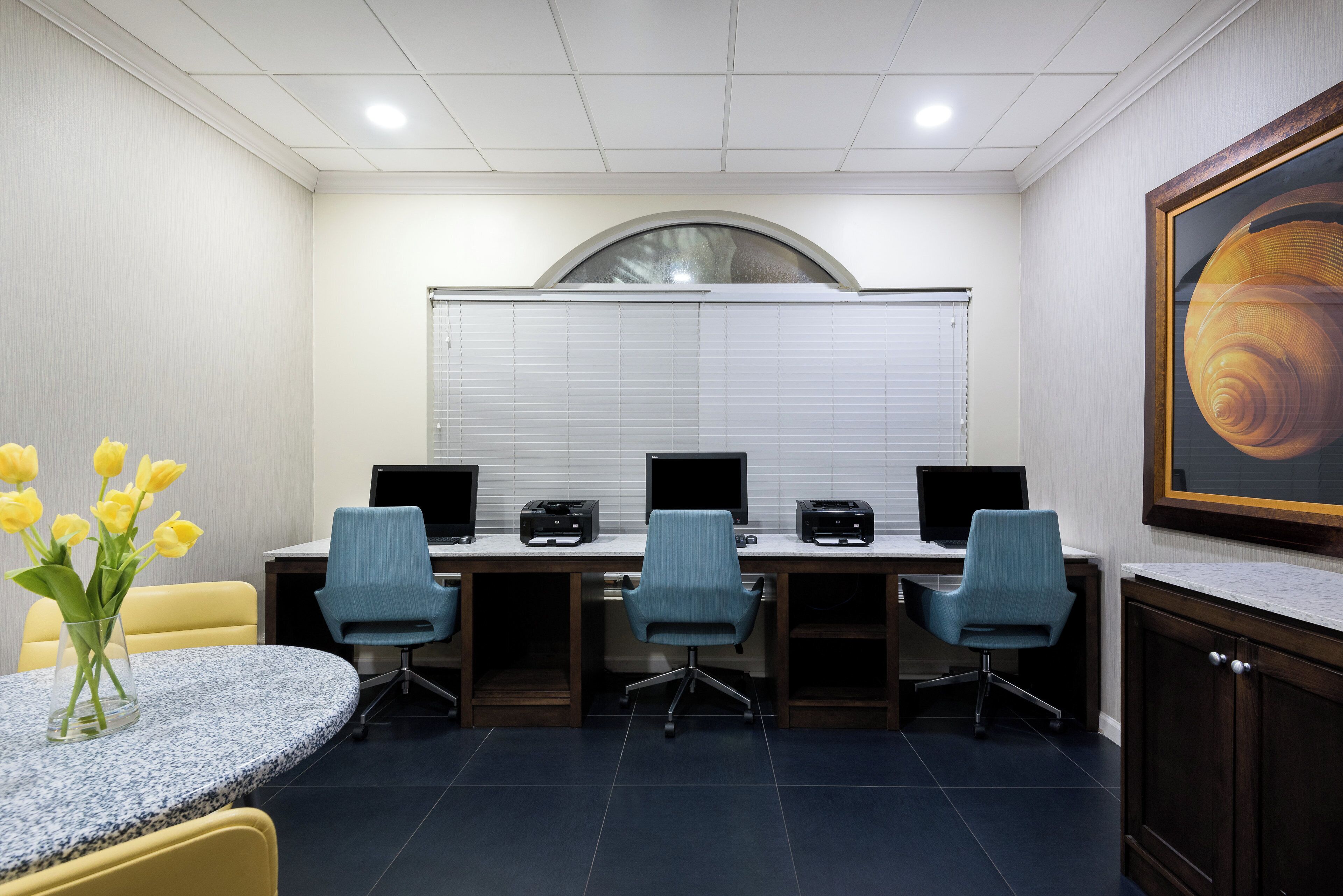 A conference room with a round table, four chairs, two desktop computers, and two printers.