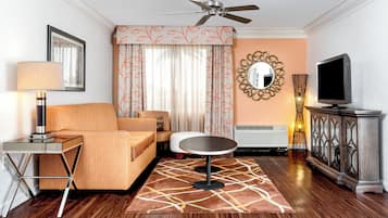 Habitación de hotel con sofá, una mesita redonda, televisor, ventilador de techo, espejo y lámpara decorativa.