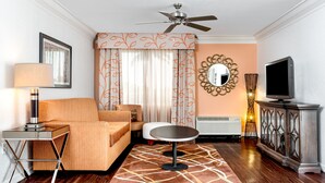 Ein Hotelzimmer mit einer Couch, einem kleinen runden Tisch, einem Fernseher, einem Deckenventilator, einem Spiegel und einem dekorativen Teppich.