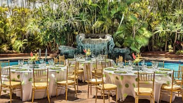 An outdoor dining area with tables set for a meal, surrounded by lush tropical vegetation and a pool.
