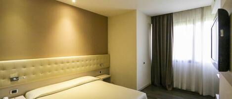 A hotel room with a large bed, a bedside table, a flat-screen TV mounted on the wall, and a window with curtains.