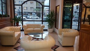 A lobby with a sofa, armchairs, and a coffee table. There are potted plants and a view of the street outside.
