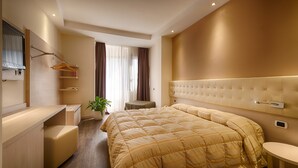 A modern hotel room with a large bed, a bedside table, a small seating area, a vanity with a mirror, and a potted plant.