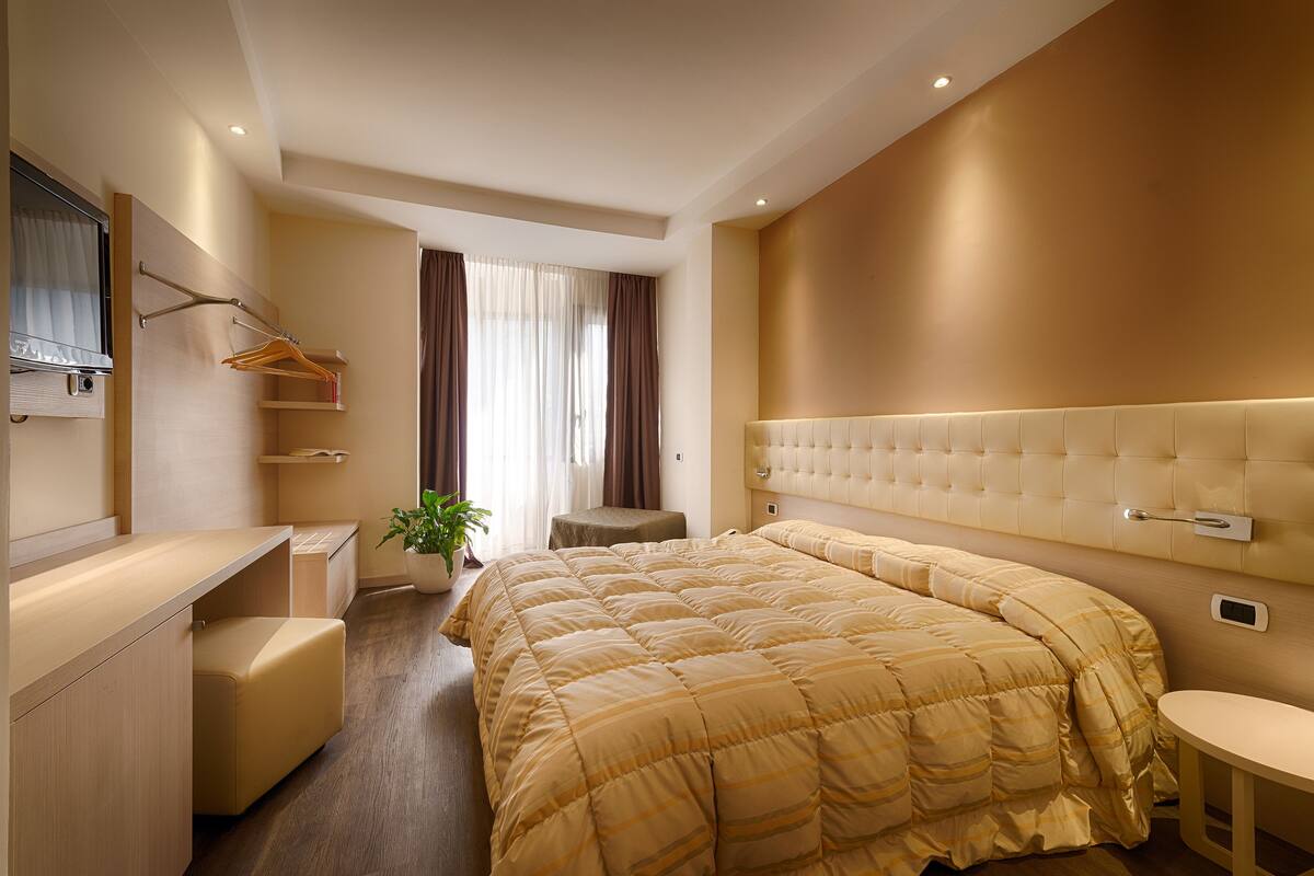 Una camera d'albergo moderna con un letto grande, un comodino, un piccolo angolo salotto, un tavolino da trucco con specchio e una pianta in vaso.