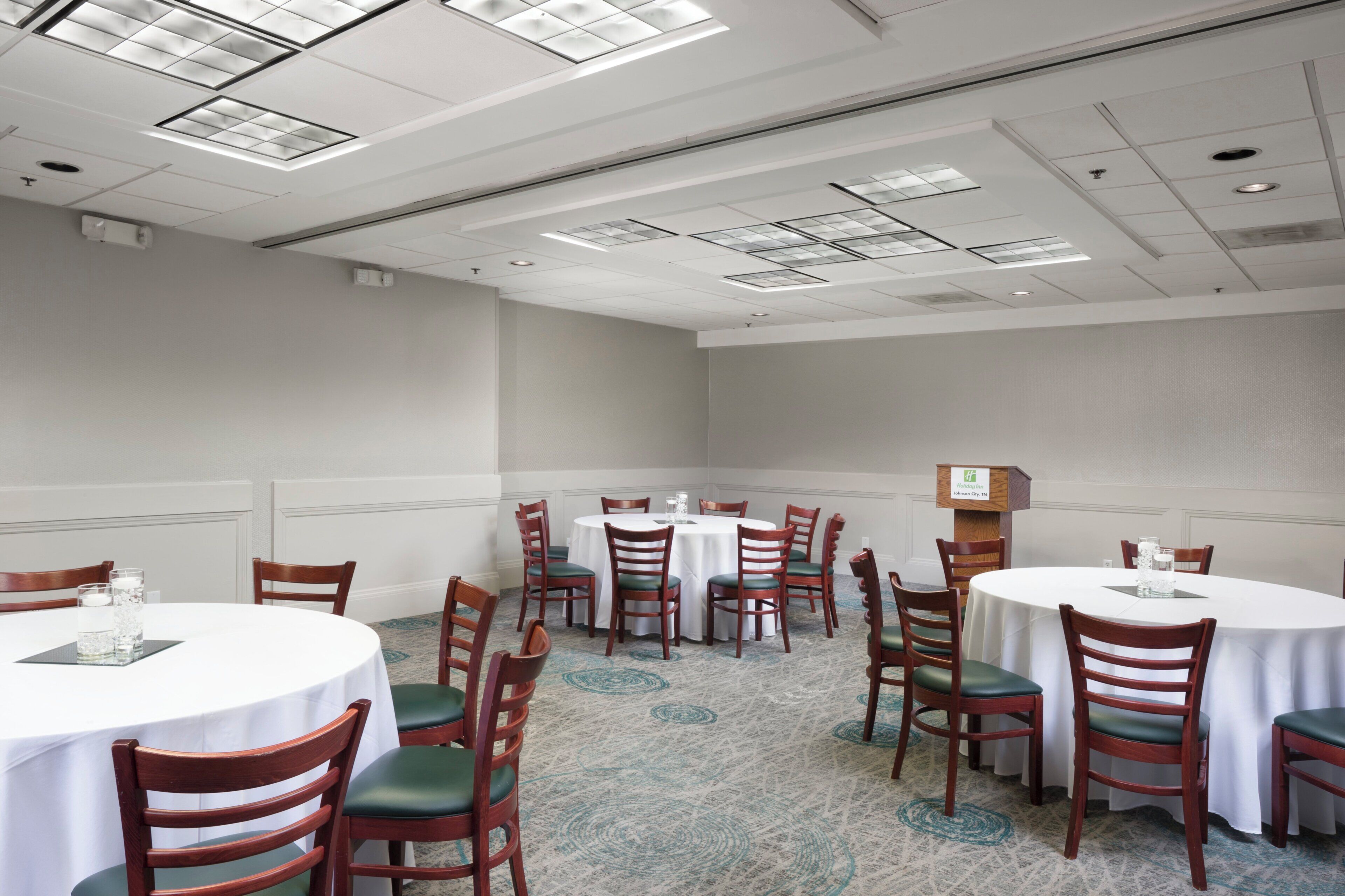 A hotel conference room with multiple round tables and chairs, a white ceiling with recessed lighting, and a carpeted floor.