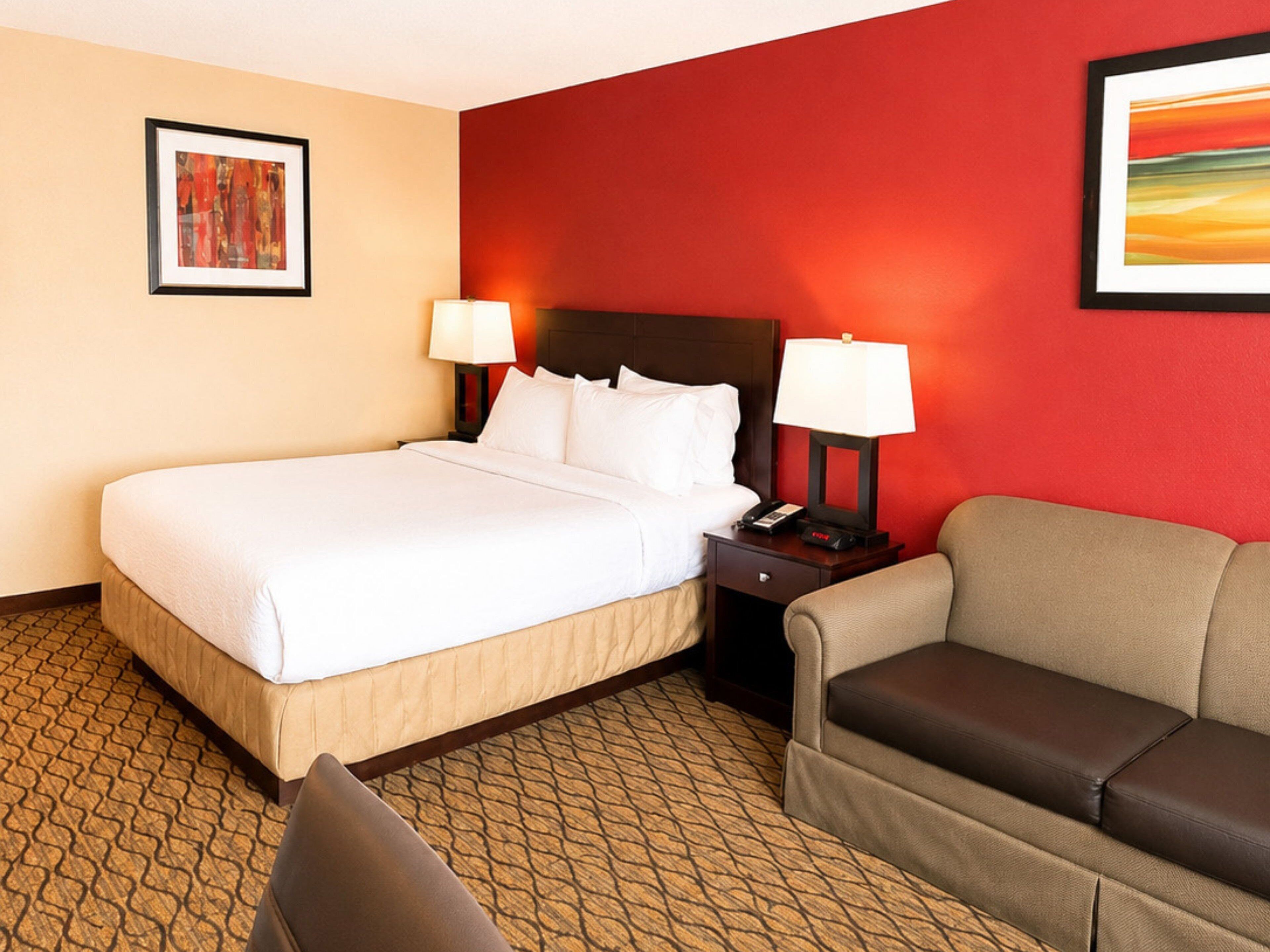 A hotel room with a bed, two bedside lamps, a nightstand, a sofa, and a patterned carpet.