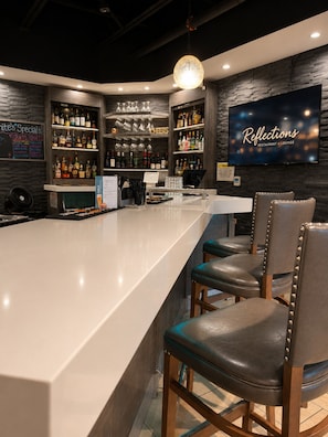 A bar area with a long counter, bar stools, a TV screen, and shelves with bottles and glasses.