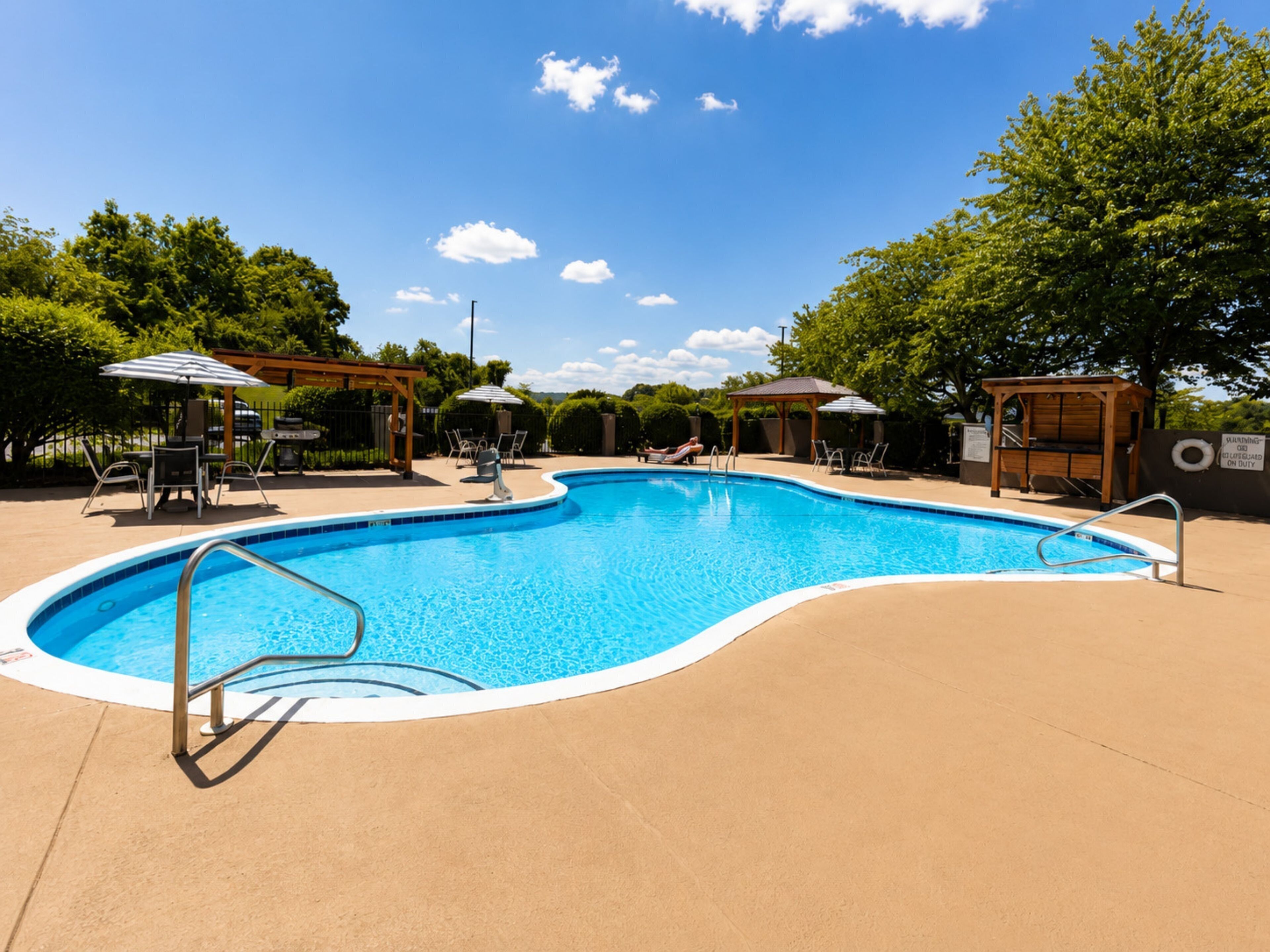 A swimming pool with a curved edge, surrounded by lounge chairs and umbrellas, with trees and a clear sky in the background.