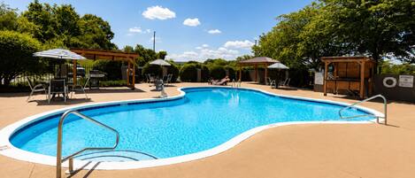 A swimming pool with a curved edge, surrounded by lounge chairs and umbrellas, with trees and a clear sky in the background.