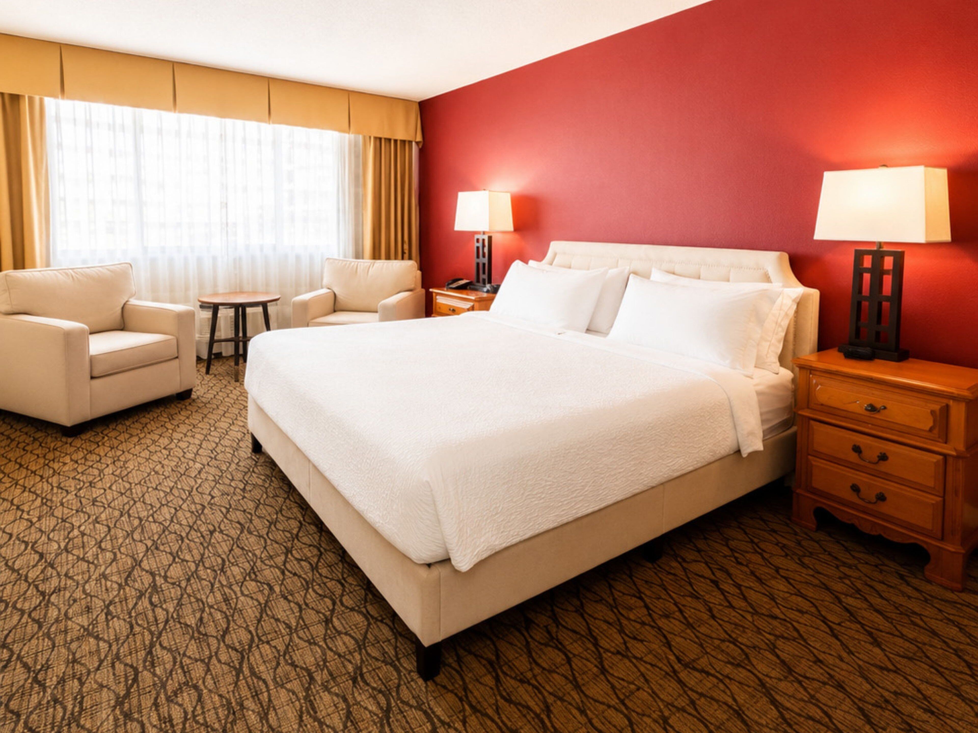 A hotel room with a large bed, two bedside tables, a chair, a small table, and a window with curtains.