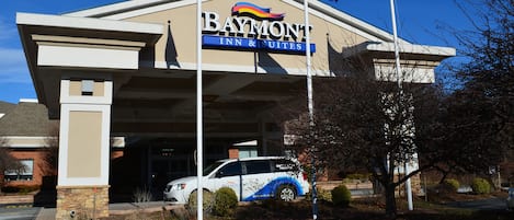 Edificio de Baymont Inn & Suites con fachada blanca y azul, un asta con la bandera estadounidense y un SUV blanco estacionado.