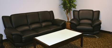 A room with a black leather sofa, a matching armchair, and a white coffee table. There is a patterned carpet and a potted plant in the corner.
