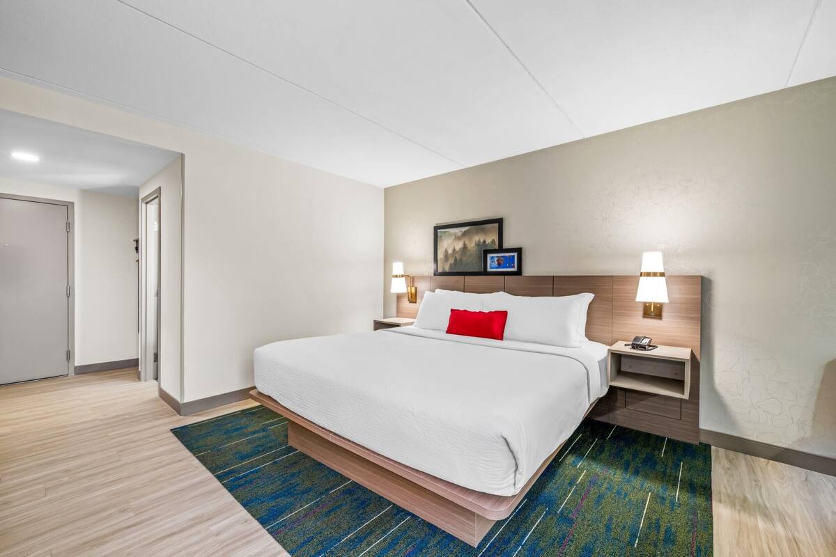 Una habitación de hotel con una cama grande, dos mesitas de noche, una mesita auxiliar, una lámpara de pared y una puerta.