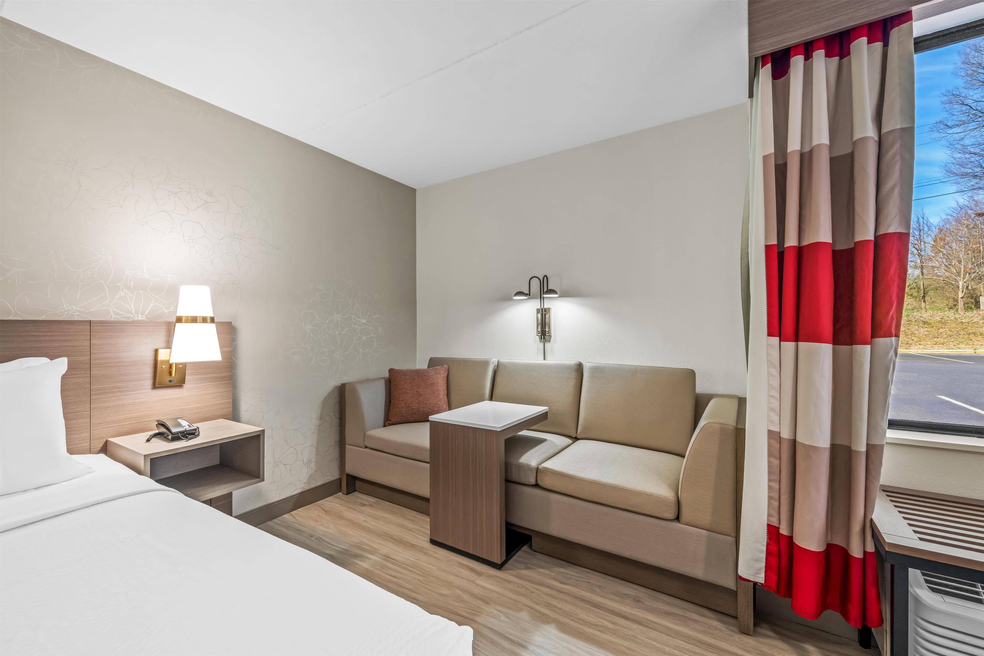 Habitación de hotel con cama, sofá, mesita de noche con lámpara, luz de pared y ventana con cortinas.