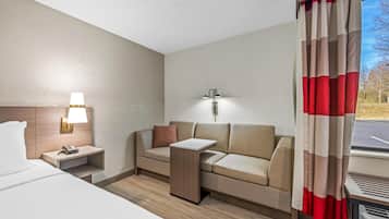 Habitación de hotel con cama, sofá, mesita de noche con lámpara, luz de pared y ventana con cortinas.